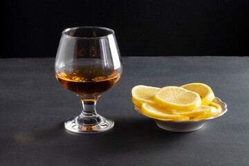 On the table is a glass of cognac. Nearby is a plate of sliced lemons.