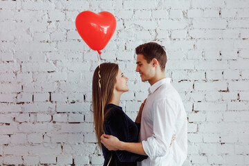 Obraz na płótnie Canvas Beautiful young couple hugging on the white wall background while celebrating Saint Valentine's Day with air balloon in shape of heart in hands.
