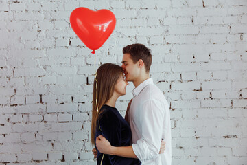 Fototapeta na wymiar Love is in the air. Attractive young couple hugging and kissing on the white wall background while celebrating Saint Valentine's Day with air balloon in shape of heart in hands.