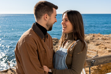 Optimistic young couple laughing and looking at each other while standing on terrace during Saint Valentine Day celebration on rocky shore of sea