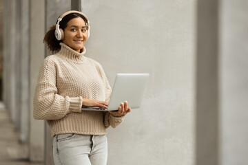 A young afro american woman standing in an urban environment with wireless headphones on holding a laptop.