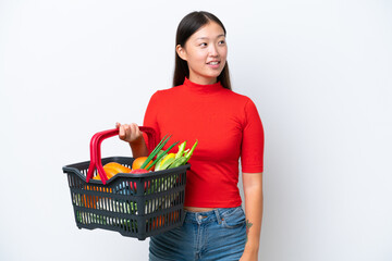 Fototapeta na wymiar Young Asian woman holding a shopping basket full of food isolated on white background looking side