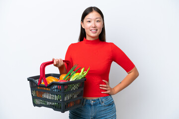 Young Asian woman holding a shopping basket full of food isolated on white background posing with arms at hip and smiling