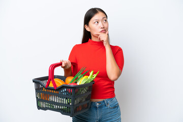 Obraz na płótnie Canvas Young Asian woman holding a shopping basket full of food isolated on white background having doubts