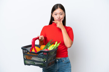 Fototapeta na wymiar Young Asian woman holding a shopping basket full of food isolated on white background thinking