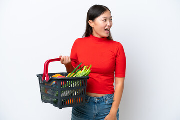Young Asian woman holding a shopping basket full of food isolated on white background laughing in lateral position