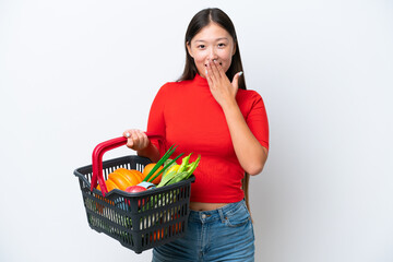 Fototapeta na wymiar Young Asian woman holding a shopping basket full of food isolated on white background happy and smiling covering mouth with hand