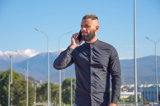 Man With A Beard And Slicked Back Hair In Black Shirt Talking On Phone, Stands On The Street Of The City From Behind Against The Background Of Lanterns, Pillars And Highlands Of The Mountains.