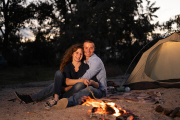 Man and woman tourists sitting in front of tent at campfire and forest background. Night camping on shore. Active outdoor lifestyle