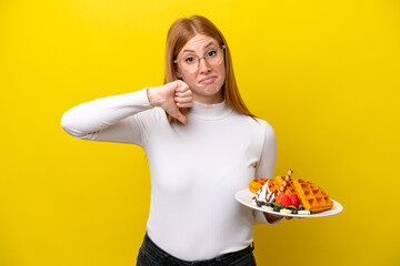 Young redhead woman holding waffles isolated on yellow background showing thumb down with negative expression
