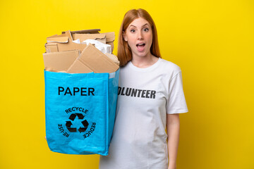 Young redhead woman holding a recycling bag full of paper to recycle isolated on yellow background with surprise facial expression