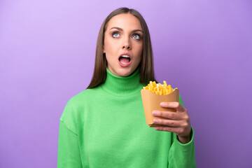 Young caucasian woman holding fried chips on purple background looking up and with surprised expression