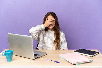 Little student girl in a workplace with a laptop isolated on purple background covering eyes by hands and smiling