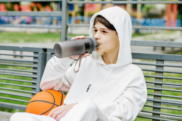 portrait of teenager boy sitting on bench with basketball and drinking water during break