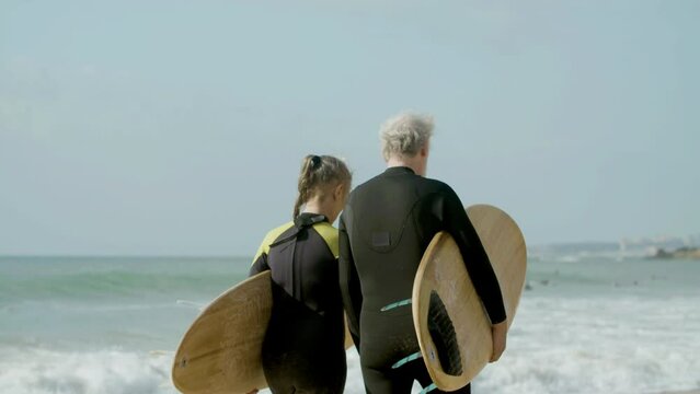 Elderly couple with surfboard walking along ocean shore. Back view of man and woman in wetsuit getting ready for surfing, spending time together. Family, retirement, extreme sport concept