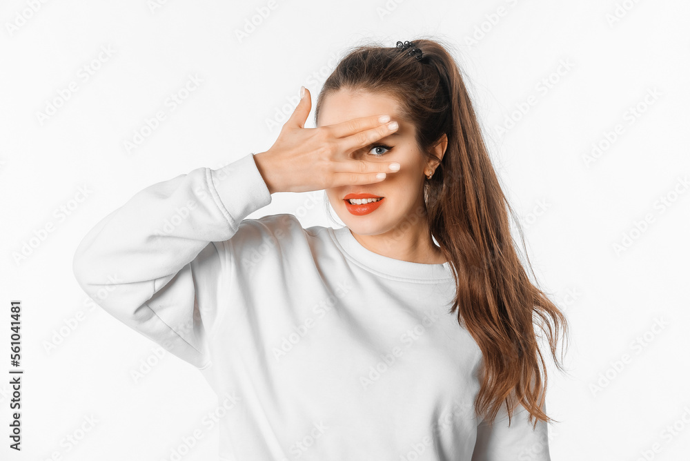 Wall mural Shocked, worried woman, hiding face behind hands, peeking through fingers surprised, looking with a happy smile on face, standing against white studio background - Wall murals