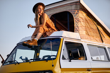 Dreamy calm female with long hair sits on roof of yellow van for trip, enjoy, having rest alone, in countryside in rural nature. holidays at summer, sunset. tourism, travel, people lifestyle concept