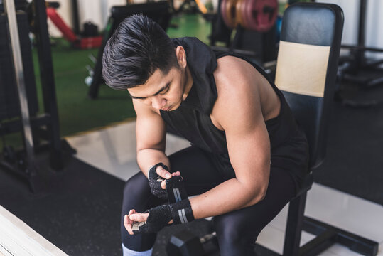 An asian guy fixes his weightlifting gloves while sitting down on the bench. Working out at the gym.