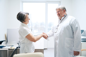 Fototapeta premium Friendly doctor greets with handshake elderly woman in medical office