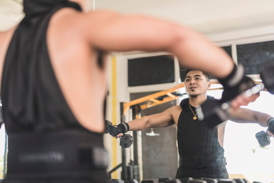 A fit asian guy performing a set of dumbbell lateral raises. Shoulder workout at the gym. Wearing a black low cut tank top and a weightlifting belt.