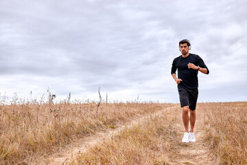 Strong and fit caucasian sportsman is running forward across the field at summer morning, alone. Active workout on fresh air before marathon. Recreation, healthy lifestyle, sport, fitness concept.