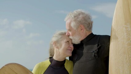 Senior surfers in wetsuits hugging in background of ocean. Close-up shot of peaceful wife and husband with surfboards in hands embracing, looking and posing for camera. Love, sport, retirement concept