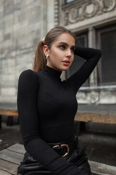 Beautiful fashionable elegant fresh girl model with earrings in trendy black clothes sits in the city. Urban female portrait of pretty woman