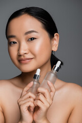 pleased asian woman with naked shoulders holding bottles with cosmetic serum and looking at camera isolated on grey.