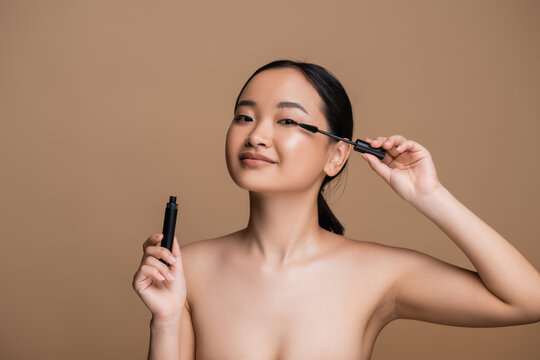 Young asian woman with naked shoulders holding mascara isolated on brown.