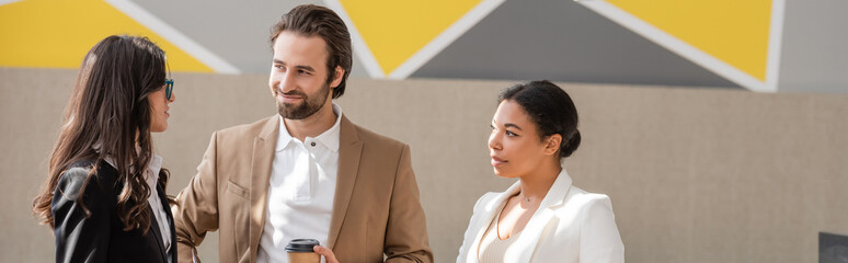 smiling manager with takeaway drink looking at colleague near multiracial businesswoman in office, banner.