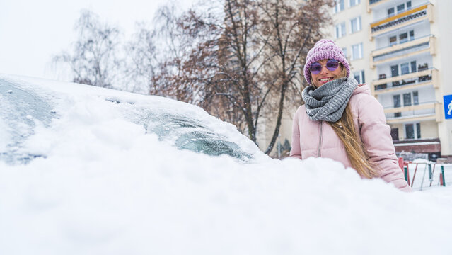 Happy young caucasian woman in pink jacked cleaning irst snow from her car. High quality photo
