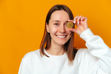 Cheerful woman hides one eye behind a golden bitcoin coin while smiling.