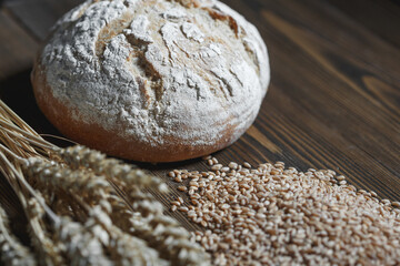 Fresh baked wheat bread loaf, a ghandful of wheat grain and wheat ears on a dark brown wooden background.