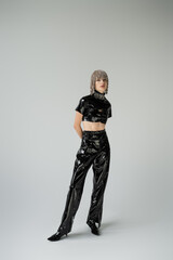 Full length of stylish woman in jewelry headwear and latex clothes standing on grey background.