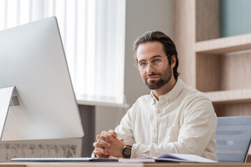 young bearded businessman in eyeglasses sitting with clenched hands near computer monitor and...