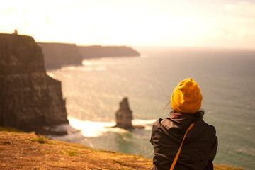 Girl looks at the ocean from behind. In front of the very high cliffs of Ireland. Warm colors at sunset.