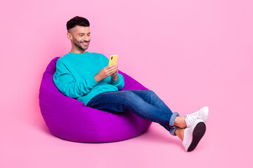 Full body photo of young handsome man sit bean bag reading blog hold gadget dressed stylish blue look isolated on pink color background