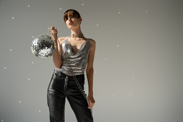 Naklejka premium stylish woman in shiny top and leather pants holding disco ball on grey background.