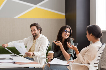 positive businesswoman in eyeglasses holding paper cup and talking to multiethnic colleague in office.