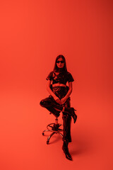 full length of brunette woman in latex crop top and trousers sitting on stool while looking at camera on red.