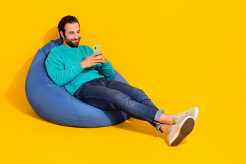 Full length photo of dreamy positive guy teal outfit sitting bean bag communicating gadget empty...