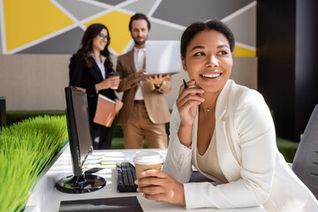 happy multiracial woman sitting with coffee to go near computer and colleagues on blurred background.