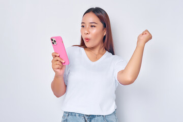 Beautiful excited Asian girl wearing casual white t-shirt holding mobile phone, raising her fist, celebrating good luck isolated on white background. People lifestyle concept