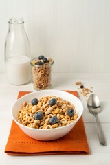 Cereal bowl, milk bottle and spoon on an orange napkin on a white table, front view.