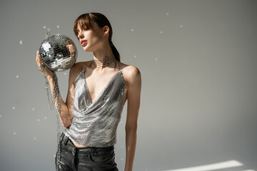 young woman in trendy top and leather pants holding chain with disco ball on grey.