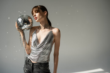 Naklejka premium young woman in trendy top and leather pants holding chain with disco ball on grey.