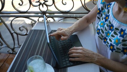 Closeup view of mature senior womans hands using a laptop tablet computer with a drink on a table at cafe on balcony in europe. Traveling digital nomad concept.