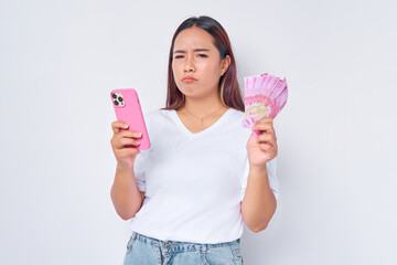 Sad young blonde woman girl Asian wearing casual white t-shirt using mobile phone and holding money rupiah banknotes isolated on white background. Financial investment concept