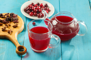 A teapot and a cup of hibiscus tea next to the table on a wooden board anise and cinnamon and hibiscus flowers and cherries in a plate.