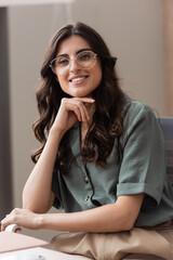brunette businesswoman in eyeglasses holding hand near chin while smiling at camera in office.